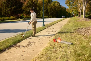 A person using a string trimmer to edge a sidewalk in a suburban area, with a leaf blower on the grass.