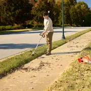 A person using a string trimmer to edge a sidewalk in a suburban area, with a leaf blower on the grass.