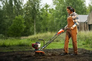 Husqvarna T 300RH Compact Pro. Person, die bei Tageslicht eine Motorhacke auf einem Stück Erde in der Nähe eines Holzhauses bedient.