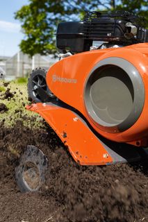 Eine Husqvarna Motorhacke in Aktion, die den Boden in einer Gartenumgebung aufwühlt.
