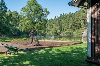 En person som använder en jordfräs för att förbereda jord i en trädgård nära en sjö.