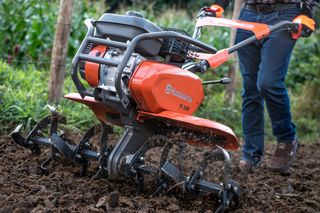 A person using a Husqvarna tiller to cultivate soil in a garden.