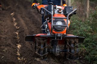 A person operating a Husqvarna tiller on a muddy field.