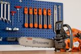 Uma motosserra Husqvarna colocada sobre uma mesa de madeira com várias ferramentas e acessórios pendurados num suporte de parede.