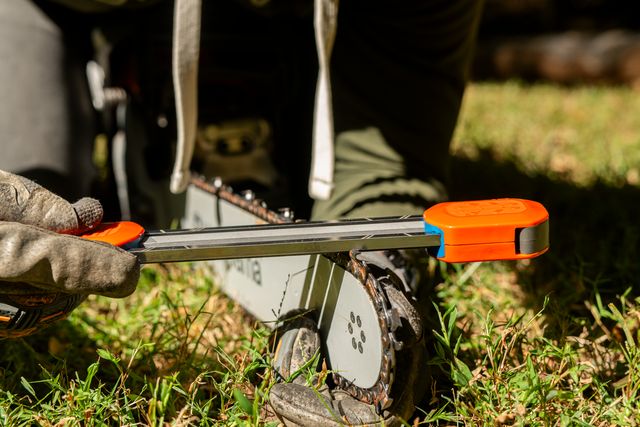 Una persona afilando la cadena de una motosierra con una herramienta de guía de afilado sobre una superficie cubierta de hierba.