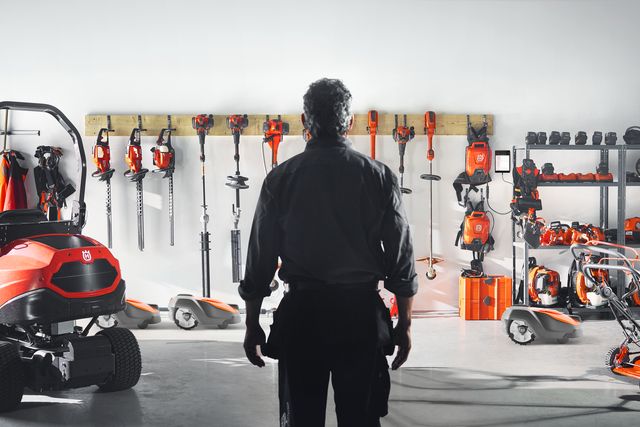 A person standing in a garage with various Husqvarna gardening and construction tools displayed on the wall.