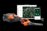 Husqvarna robotic lawn mower, leaf blower, and remote control displayed with a laptop and smartphone showing a garden planning app.