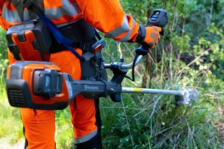 En person i arbetskläder som använder en Husqvarna buskröjare i trädgårdsmiljö.