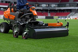 Une personne utilisant une tondeuse autoportée Husqvarna avec un grand accessoire sur un terrain de sport.