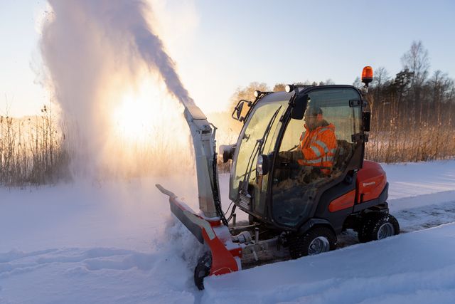 En person som kör ett snöslungefordon som röjer snö på en väg under soluppgången.
