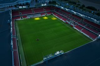 Vue aérienne d’un stade de football bien éclairé avec de l’herbe verte et des sièges rouges.