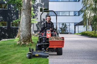 En person som kör en åkgräsklippare, klipper gräs nära ett träd och en modern byggnad.