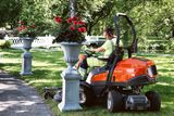 Une personne tond la pelouse avec une tondeuse autoportée dans un jardin, entourée de pots de fleurs.