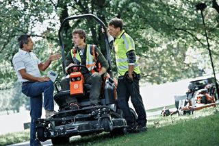 Three people discussing near a ride-on mower in a park setting.