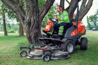 A person operating a ride-on mower near a large tree in a grassy area.