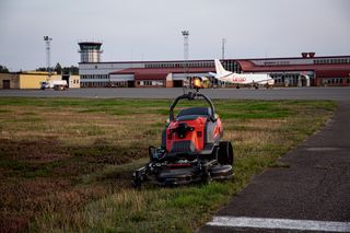 Fjärrstyrd gräsklippare som arbetar på ett flygfält nära en landningsbana med ett flygplan i bakgrunden.