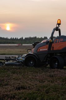 Egy Husqvarna kereskedelmi forgalomban kapható elülső fűnyíró, amely naplementekor füves területen működik.