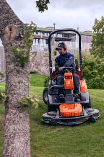 Una persona che taglia il prato con un trattorino tagliaerba in un giardino, un'altra persona che utilizza un soffiatore sullo sfondo.