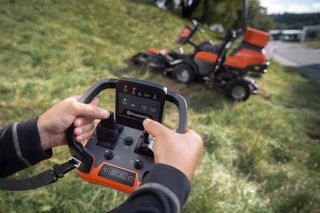 En person som använder en fjärrkontroll för att styra en professionell frontrotorklippare från Husqvarna i en gräsbevuxen sluttning.