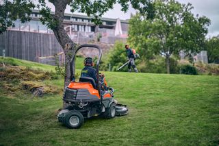 Une personne tond une pelouse avec une tondeuse autoportée pendant qu’une autre personne utilise un coupe-bordure dans un jardin.