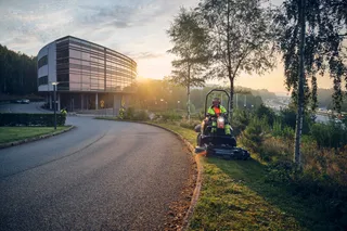 A person operating a ride-on mower on a grassy area near a modern building at sunrise.