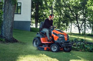 A person mowing lawn with a ride-on mower near a house and trees.