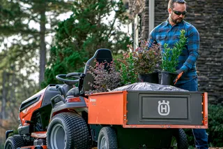 A person transporting plants and soil in a trailer attached to a ride-on mower in a garden.