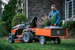 A person loading plants and a bag into a Husqvarna ride-on mower trailer in a garden.