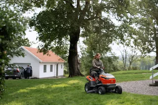 A person mowing a lawn with a ride-on mower near a house in a garden setting.