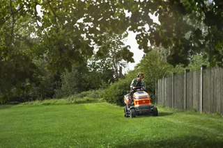 Een persoon die een gazon maait met een zitmaaier naast een houten hek in een tuin.