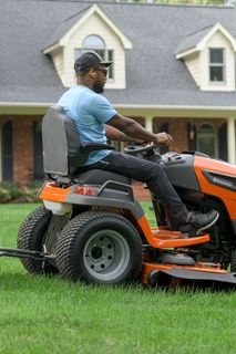 A person mowing lawn with a ride-on mower in front of a large house.