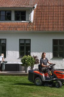 Eine Person, die mit einem Aufsitzmäher den Rasen in einem Garten mäht, mit einem Haus im Hintergrund.
