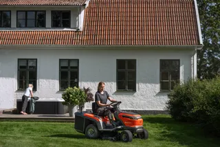 A person mowing lawn with a ride-on mower in a garden, with a house in the background.