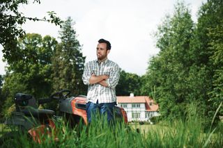 A person standing beside a ride-on mower in a garden setting with a house in the background.