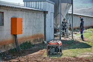 Two workers in work gear near a large industrial structure with a generator in the foreground.