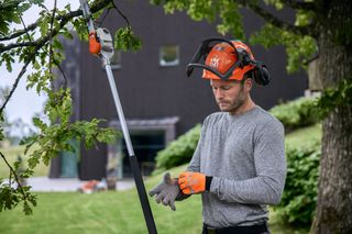A person wearing work gear and gloves, preparing to use a pole saw on a tree branch.