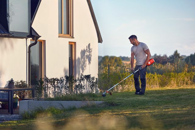 Een persoon die een grastrimmer gebruikt om het gazon in de buurt van een modern huis te onderhouden.