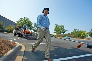 A worker in a parking lot, with a truck unloading landscaping equipment in the background.