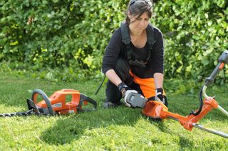 A person in work gear adjusting a battery on a garden trimmer, with a hedge trimmer beside them.