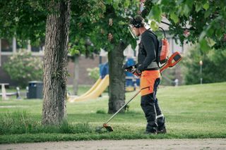 A person using a grass trimmer in a park, wearing protective work gear.