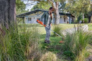 Une personne utilise un coupe-bordures pour entretenir l’herbe dans un jardin près d’une maison.