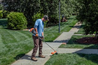 A person using a string trimmer to edge a sidewalk in a garden setting.