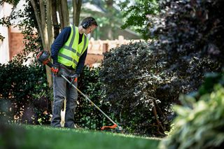 A person using a grass trimmer in a garden, wearing work gear and ear protection.