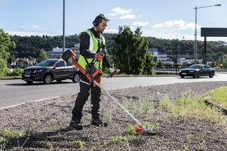 A person using a grass trimmer on a roadside area, wearing work gear.