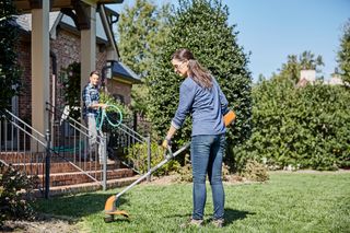A person using a grass trimmer on a lawn, with another person holding a garden hose near a house.