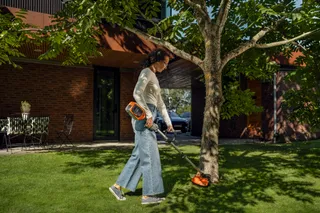 Osoba używająca podkaszarki do trawy do pielęgnacji trawnika wokół drzewa w ogrodzie.