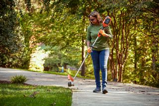 A person using a grass trimmer to edge a lawn along a sidewalk in a garden setting.