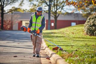 A person in work gear using a grass trimmer along a roadside curb.