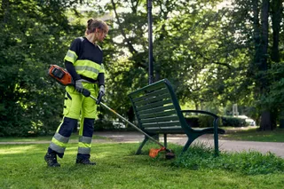 A person using a grass trimmer to cut grass around a park bench.