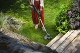 Person trimming lawn edges with a grass trimmer beside a wooden deck and garden plants.